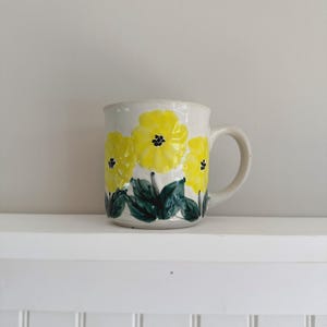 May include: A ceramic mug with a cream-coloured base featuring a hand-painted design of three bright yellow flowers with dark green leaves and black centres. The mug has a rounded handle on the right side and a smooth, glossy finish throughout.