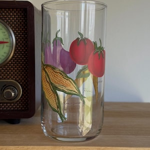 May include: A clear glass tumbler featuring hand-painted vegetable illustrations: red tomatoes, purple aubergines, and corn. The cylindrical glass is suitable for drinks. The design includes the artist's signature.