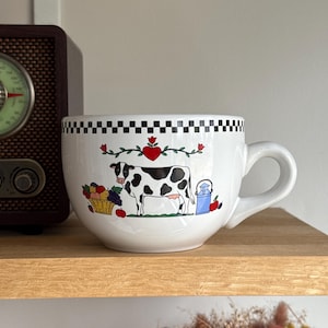 Peut inclure: Mug à café blanc en céramique surdimensionné avec une illustration de vache noire et blanche. La tasse présente une bordure à carreaux noirs et blancs, un cœur et un panier de fruits. Un pot à lait bleu et des pommes rouges sont également représentés.