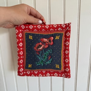 Peut inclure: Un dessous de plat matelassé carré avec un panneau central bleu foncé orné de coquelicots rouges brodés et de petites étoiles jaunes. La bordure est en tissu rouge à motif géométrique blanc avec une petite boucle de suspension.