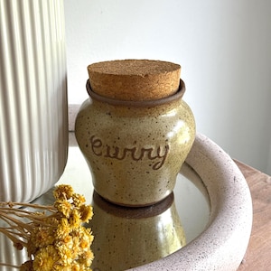 Peut inclure: Un petit pot en céramique beige moucheté avec un corps arrondi et un bord brun foncé. Le mot "curry" est gravé en écriture cursive sur le devant. Le pot est surmonté d'un couvercle rond en liège de couleur naturelle qui s'insère dans l'ouverture.