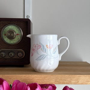 Op de afbeelding: Een witte keramische kan met een bloemenpatroon in roze, perzik en blauwgroen. De kan heeft een handvat en een tuit. Hij staat op een houten plank met een vintage radio en roze tulpen op de voorgrond.