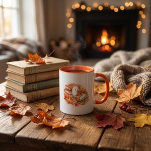Puede incluir: Una taza de cerámica blanca con borde y asa naranja, que presenta un gráfico de un pequeño fantasma blanco leyendo un libro entre una pila de libros y hojas de otoño. La taza se encuentra sobre una superficie de madera junto a cuatro libros.