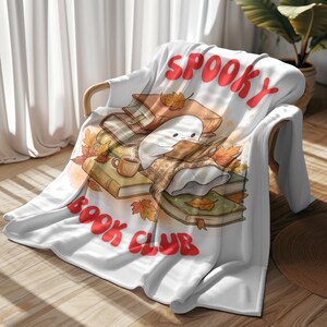 Puede incluir: Una manta blanca con una ilustración gráfica de un pequeño fantasma blanco leyendo un libro sobre una pila de libros con hojas de otoño, una taza de café y el texto rojo SPOOKY BOOK CLUB impreso en una fuente curva arriba y abajo.