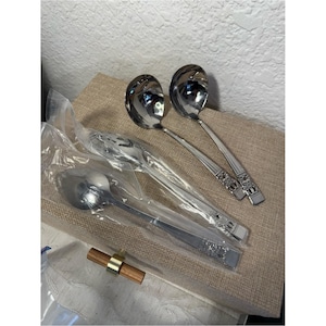 May include: Three stainless steel spoons with polished bowls and decorative handles. Two spoons are unwrapped, while one spoon remains inside a clear plastic sleeve. The handles feature a textured, embossed pattern near the base of each spoon.