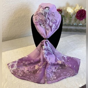 May include: A sheer, lavender scarf with a bird and floral print. The scarf is draped over a black mannequin head, showcasing its design. The bird is perched on a branch, surrounded by delicate flowers. The scarf is secured with a dark brown ring.