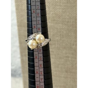 May include: A silver-toned ring featuring two faux pearls and small accent stones. The ring is on a black ring sizer. The ring size appears to be between a size 7 and 8.