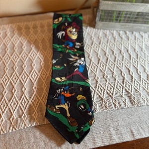 May include: Black necktie featuring cartoon characters in action poses. The tie has a black background with green accents and cartoon characters in various colors. The characters include Bugs Bunny, Daffy Duck, and Marvin the Martian.