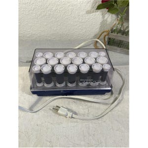 May include: A Remington Aqua Curl hair setter featuring a rectangular blue base and a clear plastic lid. Inside, multiple dark gray hair rollers with white caps are arranged in rows. A white electrical power cord with a two-prong plug is attached.