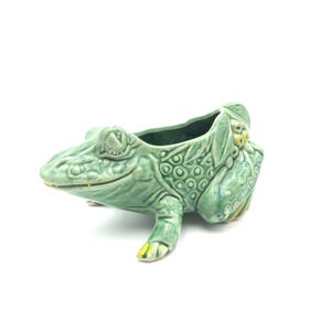 May include: A green ceramic frog planter with a detailed textured design. The frog has yellow accents on its feet and a wide open mouth. The planter is designed to hold small plants or flowers.