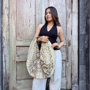 Puede incluir: Un bolso de hombro grande estilo hobo con un patrón de piel de serpiente texturizado en tonos amarillo, gris y blanco. El bolso tiene una silueta holgada y desestructurada con una correa ancha integrada y una textura de escamas visible.