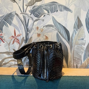 May include: Black bucket bag with a textured snakeskin pattern, a drawstring closure, and a zippered front. The bag has a shoulder strap and silver-toned hardware. The background features a floral and foliage wallpaper.