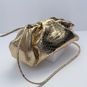 Puede incluir: Un bolso de mano en color dorado metálico con un patrón texturizado de piel de serpiente. El bolso tiene una silueta suave y fruncida tipo bolsa, con un cierre de cordón en la parte superior y una correa larga y fina a juego en los laterales.