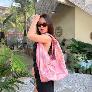 Pode incluir: Uma bolsa hobo grande e acolchoada com acabamento rosa metálico e uma textura sutil semelhante à pele de cobra. A bolsa possui uma alça larga e uma silhueta descontraída e desestruturada, projetada para ser usada no ombro.