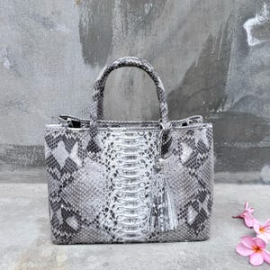 Peut inclure: Un sac à main structuré présentant un motif en peau de serpent gris et blanc avec une texture d'écailles distincte. Le sac comprend deux poignées supérieures et un pompon en peau de serpent gris assorti suspendu sur un côté.