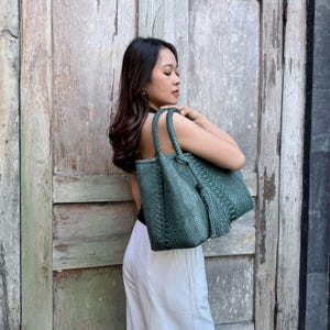 May include: A dark green tote bag with a textured, woven-like surface pattern. The bag features two sturdy top handles and a decorative tassel hanging from one side. The design is structured with a rectangular base and a spacious main compartment.