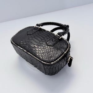 Puede incluir: Un bolso de mano pequeño y rectangular de color negro con un patrón de piel de serpiente texturizado en todo el exterior. El bolso incluye dos asas cortas trenzadas y un cierre de cremallera en el borde superior, con herrajes metálicos visibles.