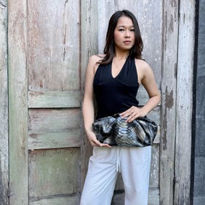 Puede incluir: Un top negro sin mangas con cuello halter, pantalones blancos y un bolso de mano tipo sobre con estampado de piel de serpiente en tonos plateado y negro, con forma redondeada y fruncida y textura suave, sostenido frente al cuerpo.