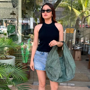 May include: A black sleeveless ribbed knit top paired with a light blue denim mini skirt and a large, textured olive green hobo bag. The top features a high neckline, and the bag has a distinct woven pattern with visible fringe details.
