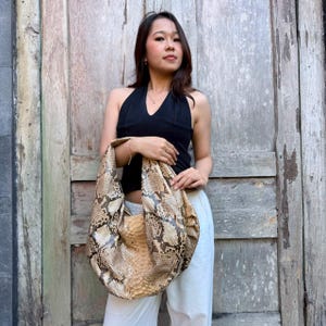 Peut inclure: Un sac à bandoulière de style hobo présentant un motif en peau de serpent texturé dans des tons beige, brun clair et brun foncé. Le sac a une silhouette affaissée en forme de croissant avec une large bandoulière intégrée et une texture d'écailles.