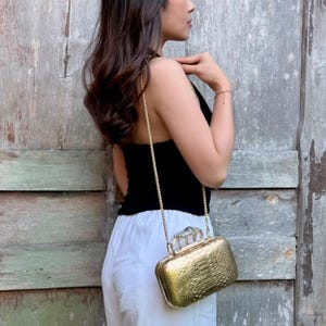 Op de afbeelding: Een metallic gouden box clutch tas met een textuur van slangenleer en een zilverkleurig handvat in de vorm van een boksbeugel met heldere kristallen accenten. De tas heeft een dunne gouden ketting als schouderband en een metalen sluiting.