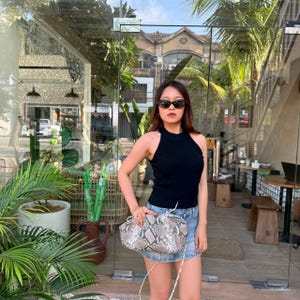 May include: A black sleeveless halter top with a ribbed texture paired with a light blue denim mini skirt and a grey and white snakeskin patterned clutch bag with a thin shoulder strap. The outfit is completed with dark oval sunglasses.