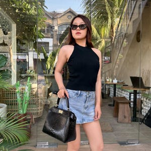 Pode incluir: Um top preto sem mangas em malha canelada combinado com uma minissaia jeans azul claro e uma bolsa preta com textura de pele de cobra e fecho de metal dourado. A bolsa apresenta uma alça superior estruturada e formato retangular.