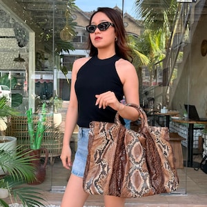 May include: A large tote bag featuring a brown and tan snakeskin pattern with a textured finish, two rounded top handles, and a spacious structured body. The bag is paired with a black sleeveless ribbed top, blue denim shorts, and black sunglasses.