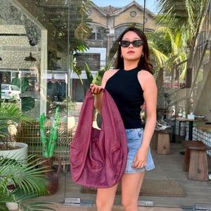 Puede incluir: Un bolso hobo de color burdeos intenso con un patrón texturizado de piel de serpiente, que presenta una silueta holgada y una correa de hombro ancha integrada. El bolso está hecho de un material flexible con un relieve de escamas uniforme.