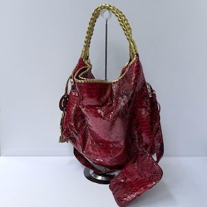 May include: A hobo-style handbag featuring a deep red and black snakeskin-patterned texture. The bag includes a metallic gold braided top handle, gold-toned trim along the edges, and a matching red snakeskin-patterned pouch attached by a strap.