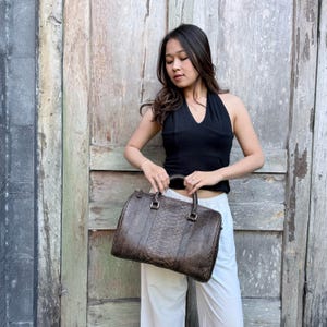 May include: A dark brown handbag featuring a textured snakeskin pattern, two top handles, and silver-toned hardware. The bag has a rectangular, structured shape with rounded corners and a zippered closure along the top edge.