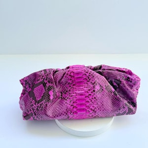 Puede incluir: Un vibrante bolso de mano magenta con un estampado de piel de serpiente. El bolso está hecho de un material suave y texturizado y tiene un diseño fruncido. El bolso de mano está sobre un soporte circular blanco.