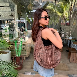 Puede incluir: Un bolso hobo con estampado de piel de serpiente marrón, silueta holgada y desestructurada con una correa ancha para el hombro, combinado con una camiseta negra sin mangas, pantalones cortos de mezclilla azul claro y gafas de sol negras.