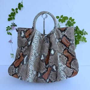 Può includere: Una borsa con motivo in pelle di serpente nei toni del marrone, beige, bianco e nero. La borsa presenta un design arricciato e plissettato con un unico manico circolare sulla parte superiore. La trama imita le squame di un serpente.