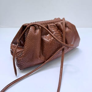 Puede incluir: Un bolso de mano marrón con un patrón texturizado de piel de serpiente, que presenta un diseño fruncido y una correa de hombro fina a juego. El bolso tiene una forma suave y redondeada con pliegues visibles y un cierre de marco estructurado.