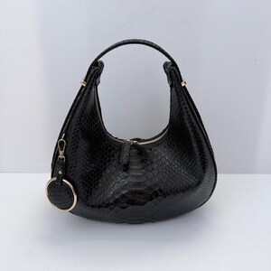 Op de afbeelding: Een zwarte hobo-tas met een gestructureerd slangenleerpatroon. De tas heeft een gebogen silhouet, een enkel handvat en een zilverkleurige ritssluiting. Een ronde bedel met een bijpassende textuur en een metalen rand hangt aan de zijkant.