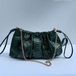 May include: A dark green, textured snakeskin-patterned handbag with a gathered, ruched design. The bag features a thin, gold-toned metal chain strap attached to the top corners with lobster claw clasps and two matching side straps.