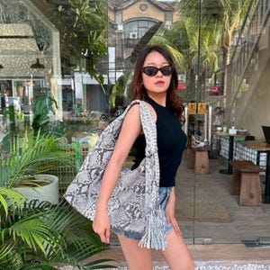May include: A large hobo-style shoulder bag featuring a grey and white snakeskin pattern with a textured finish. The bag has a wide, braided shoulder strap and a long, decorative fringe tassel hanging from the side, crafted from the same material.