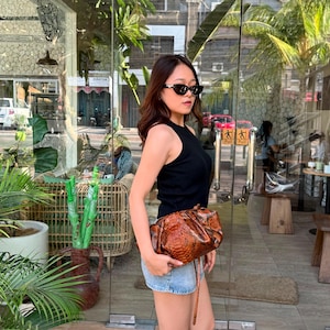 May include: A black sleeveless ribbed top, light blue denim shorts, and a brown snakeskin-patterned clutch bag. The ensemble is paired with dark-tinted cat-eye sunglasses, creating a casual look with a textured accessory as the focal point.