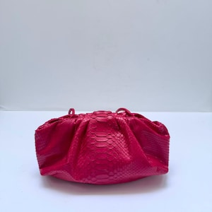 Può includere: Una pochette rosa acceso con un motivo testurizzato in pelle di serpente. La borsa presenta una silhouette arrotondata e arricciata con un pannello centrale di scaglie più grandi e scaglie più piccole sui lati, rifinita con sottili cinturini.