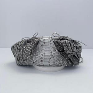 Peut inclure: Une pochette avec un motif peau de serpent gris et blanc et une finition texturée en écailles. Le sac présente une forme arrondie et froncée avec un panneau central vertical et de fines attaches à nouer sur les côtés de l'ouverture.