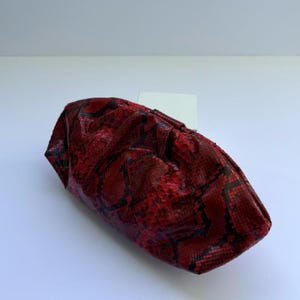 May include: A red and black snakeskin-patterned clutch bag. The bag is made of a textured material and has a gathered design. The clutch is a deep red color with black accents, and it sits on a white surface.