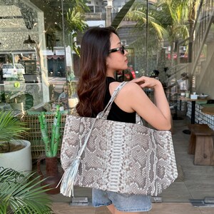 Op de afbeelding: Een grote tote bag met een slangenleerpatroon in beige, bruine en crèmetinten. De tas heeft twee schouderbanden en een lange, metallic zilveren kwast aan één kant bevestigd. De buitenkant heeft een textuur met een reptielachtig patroon.