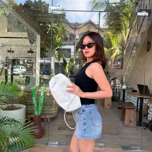 Könnte beinhalten: Ein schwarzes ärmelloses Rippstrick-Oberteil kombiniert mit hellblauen Jeansshorts und einer weißen, strukturierten Handtasche. Das Outfit wird durch eine schwarze Cat-Eye-Sonnenbrille ergänzt und besticht durch einen lässigen, schlichten Look.