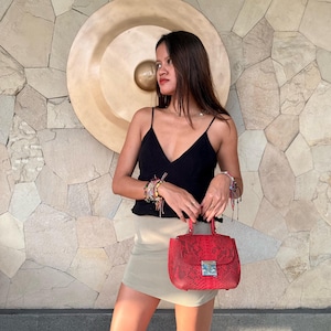 Puede incluir: Un top negro sin mangas con tirantes finos y escote en V, combinado con una minifalda beige claro y un bolso rojo con estampado de piel de serpiente, cierre metálico plateado y asa superior, sostenido frente al cuerpo.