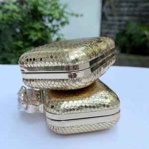 May include: Two rectangular gold-toned clutch bags with a textured snakeskin-like pattern. Each clutch features a metallic frame and a smooth, polished edge. The bags are stacked vertically, showcasing their compact, boxy shape and metallic finish.