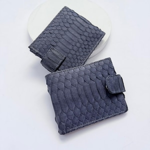 Puede incluir: Dos carteras cuadradas con una textura de piel de serpiente gris oscuro y un patrón de escamas distintivo. Cada cartera incluye una pequeña lengüeta de cierre a juego en el lateral, mostrando un diseño uniforme de inspiración reptiliana.