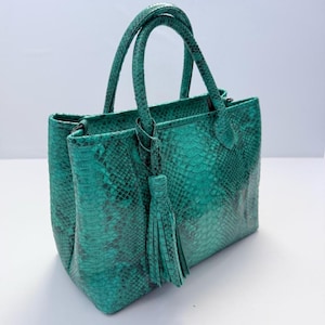 May include: A structured handbag featuring a vibrant teal and black snakeskin pattern. The bag includes two top handles and a decorative matching tassel attached to one handle. The exterior displays a detailed scale texture throughout the surface.
