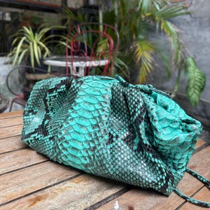 Puede incluir: Un bolso de mano de piel de serpiente turquesa y negro con cierre de cordón. El bolso está hecho de piel de serpiente auténtica y tiene un aspecto texturizado y escamoso. El bolso está sobre una superficie de madera.