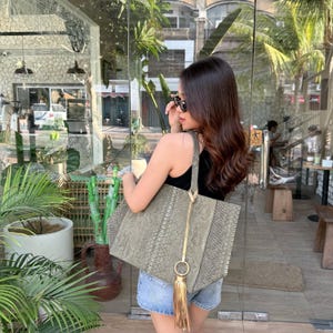 May include: A large, rectangular tote bag featuring a textured, grey-toned snakeskin pattern. The bag includes a long, thin shoulder strap and a prominent gold-toned metal ring accent attached to a long, matching gold-toned leather tassel.